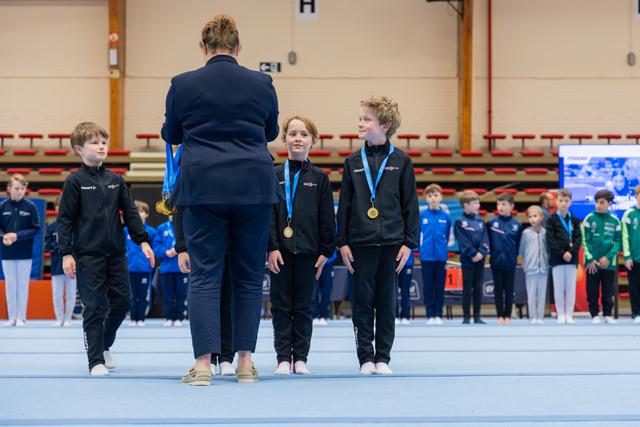 An adult presenter awards gold medals to two proud children in black Craft jackets at an indoor gymnastics event.