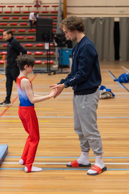 A young gymnast in red and blue receives guidance from a coach, both focused, sharing a supportive hand-to-hand moment on the gym floor.