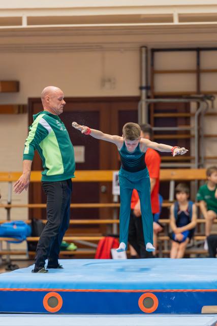 Bald coach in green jacket steadies a focused young gymnast mid-air above a blue trampoline mat indoors.