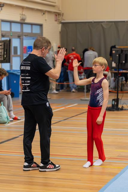 A coach and young male gymnast in red leotard share a high-five on the gym floor, showing mutual encouragement.