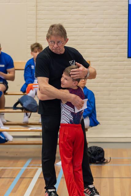 An older coach in black embraces a young gymnast in red pants, offering reassurance in a sports hall.