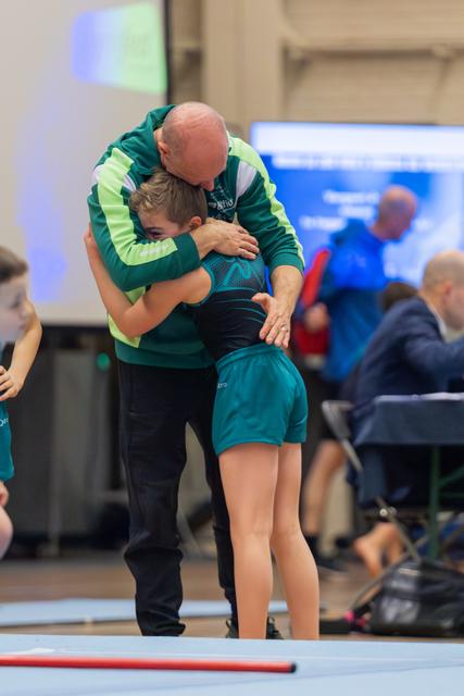 Bald coach in green jacket embraces young gymnast in teal leotard on floor mat, a moment of comfort or celebration.