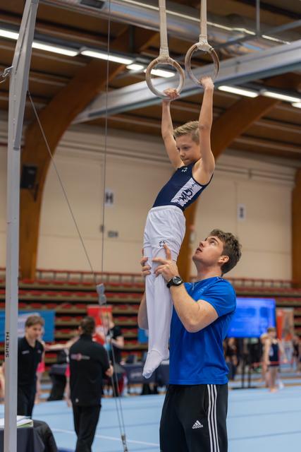 A coach steadies a focused young gymnast on still rings during a gymnastics competition indoors.