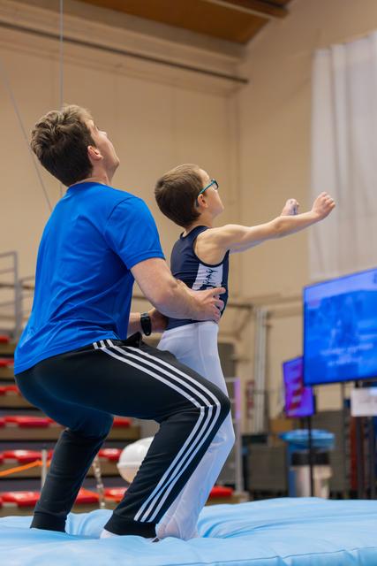 Coach steadies a young gymnast in glasses as they practice a skill together on a mat in an indoor gym.