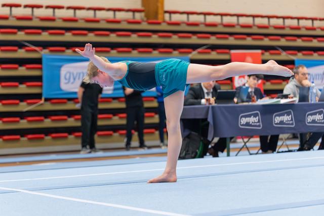 Young gymnast performs a precise arabesque on the floor mat, arms raised gracefully, judges scoring at the table behind.