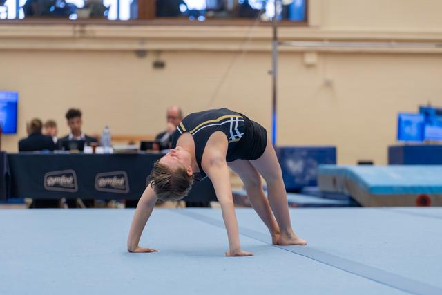 Young gymnast performing a bridge pose on the blue floor mat, head tilted back, during a Gymfed competition.