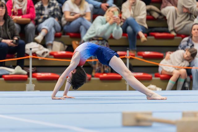 Young gymnast performs a backbend bridge on the floor exercise mat, wearing a blue and pink leotard, audience watching.