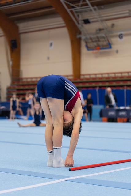 Young gymnast bending deeply forward on blue floor mat, stretching near a red gymnastics bar in a sports hall.