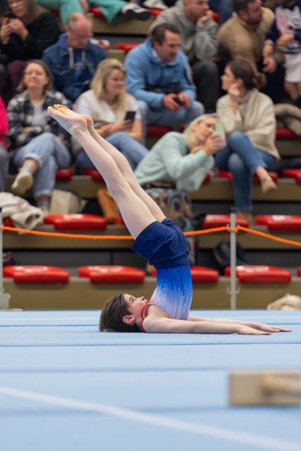 Young gymnast performs a shoulder stand on the floor exercise mat, legs extended skyward, audience watching in background.