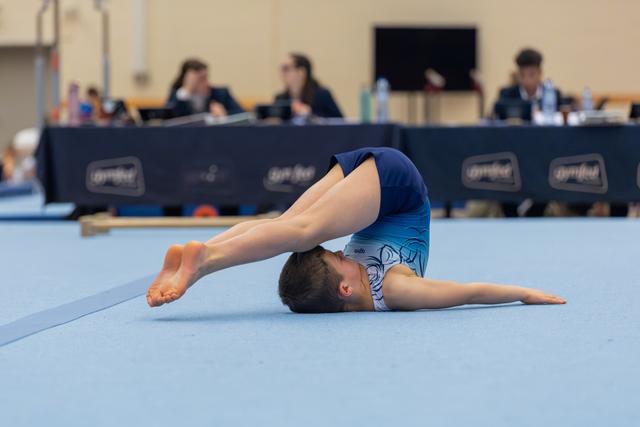 Young gymnast in blue leotard performs a tucked rollback on the floor exercise mat, judges visible in background.