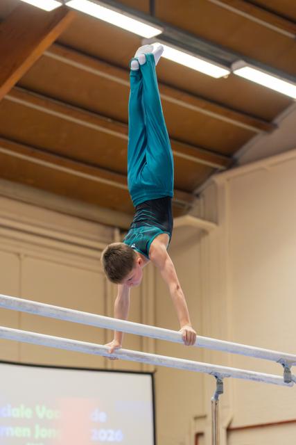 Young male gymnast performs a perfect handstand on parallel bars, legs extended upward, in an indoor gymnastics hall.