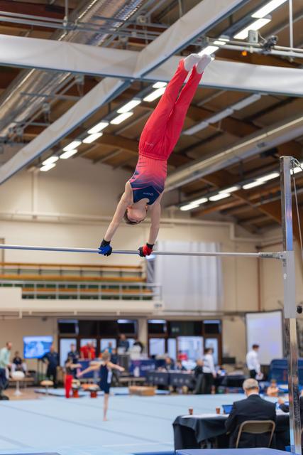 Young male gymnast in red performs a handstand on the high bar, body fully extended upside down in an indoor gymnasium.