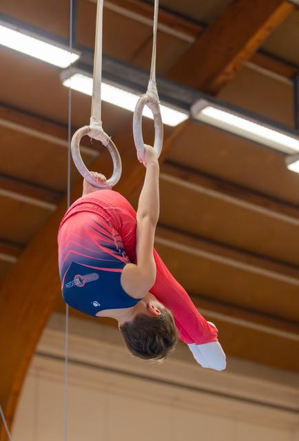 Young male gymnast hangs inverted on still rings, body curled, focused effort visible in indoor gymnasium.