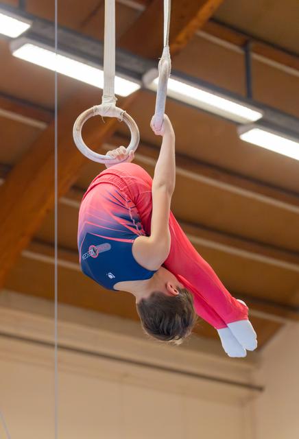 Young male gymnast hangs inverted on still rings, body arched, wearing red and blue leotard in a wooden-beamed gymnasium.