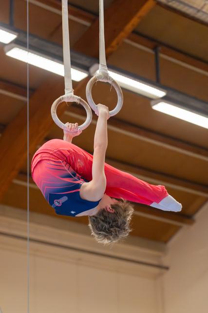 Young gymnast hangs inverted on still rings in a tuck position, wearing a red and blue leotard in an indoor gym.