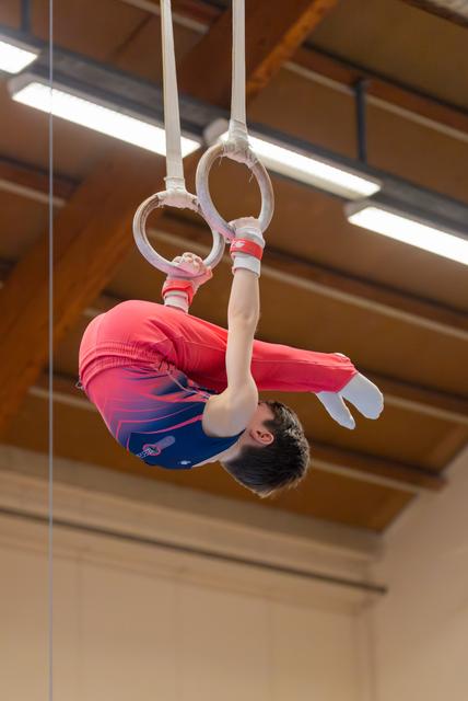 Young male gymnast performs an inverted tuck on still rings, wearing a red and blue leotard in an indoor gymnasium.
