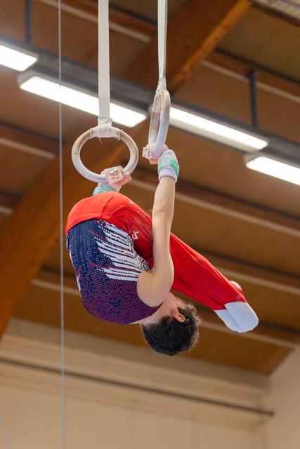 Young male gymnast performs an inverted hang on still rings, body curled in a tuck position inside a gymnasium.