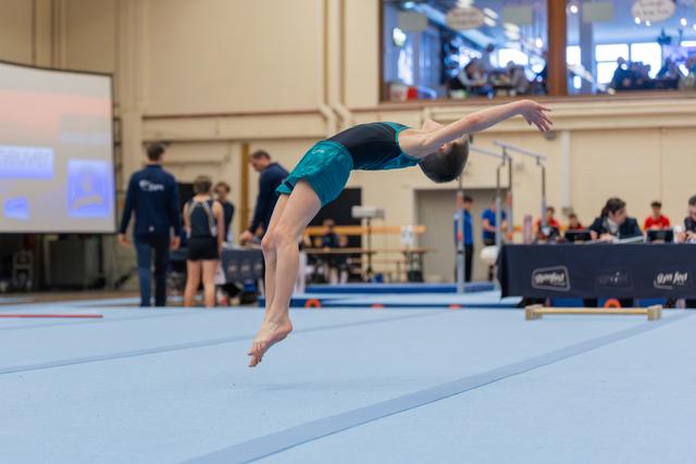 Young gymnast in teal leotard executes a powerful backflip on the floor exercise mat during an indoor competition.