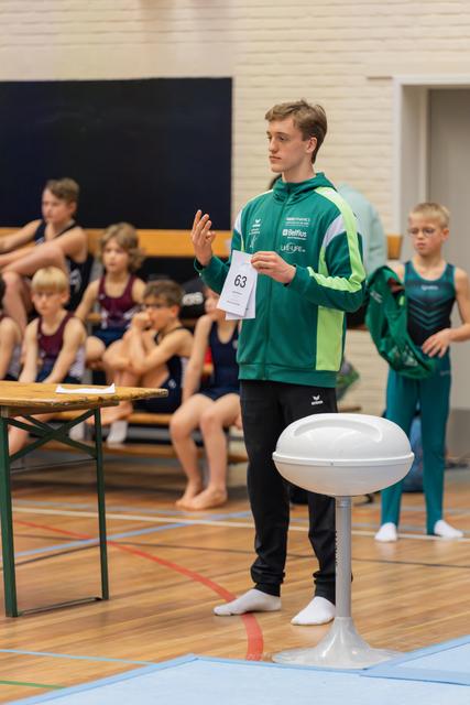Young male gymnast wearing number 63 stands confidently, gesturing while holding a score card in a gymnasium.
