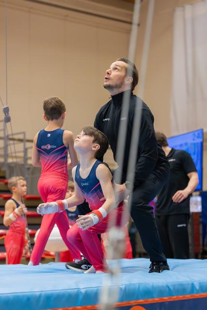 Coach spots young gymnast on mat, both gazing upward intently during a youth gymnastics meet.