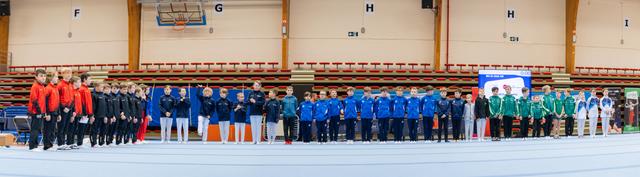 Multiple national gymnastics teams stand in a formal lineup inside a sports hall, wearing colorful team uniforms during an opening ceremony.