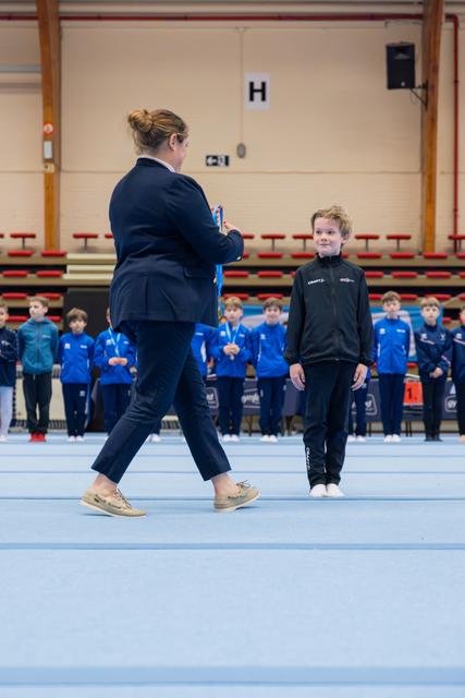 An official presents a medal to a smiling young gymnast in black, while teammates in blue line up behind them.