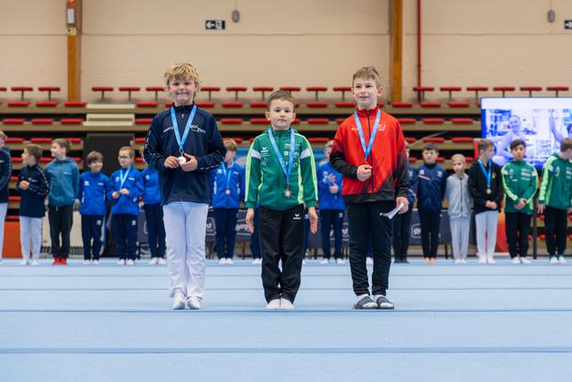 Three young boys stand proudly on the podium displaying their medals at an indoor gymnastics competition.