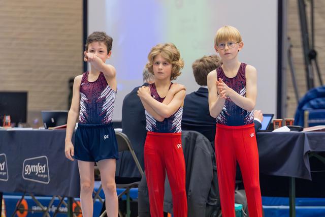 Three young gymnasts in matching uniforms watch intently, arms crossed and tense, awaiting results at a Gymfed event.