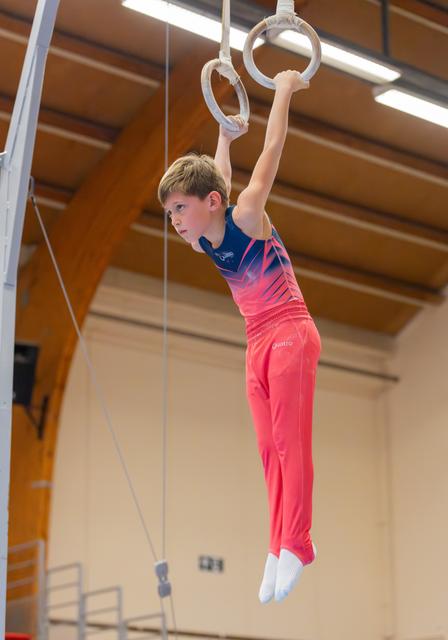 Focused young gymnast hangs from still rings in a sports hall, wearing a blue and pink leotard with red trousers.