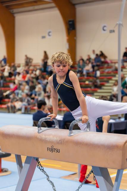Determined young gymnast grips pommel horse handles mid-routine, legs extended, concentration visible on his face. Indoor arena with spectators.