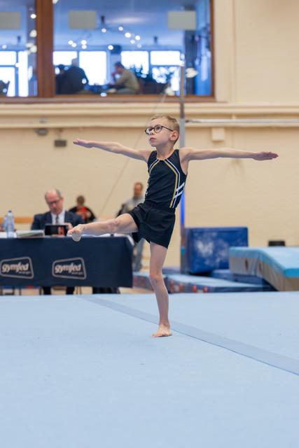 Young boy with glasses performs a precise arabesque balance on the floor exercise mat, arms outstretched with focused concentration.