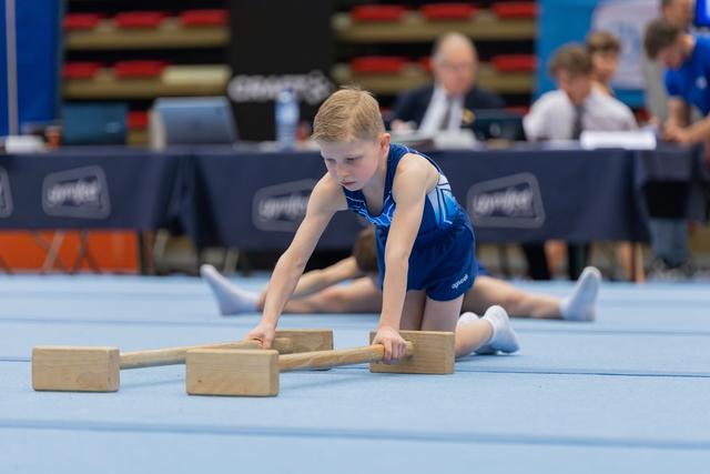 Focused young boy in blue leotard performs floor exercise with wooden apparatus blocks at a gymnastics meet.