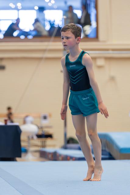 Young boy in teal Quatro gymnastics outfit stands composed on floor mat, focused expression, preparing for routine.