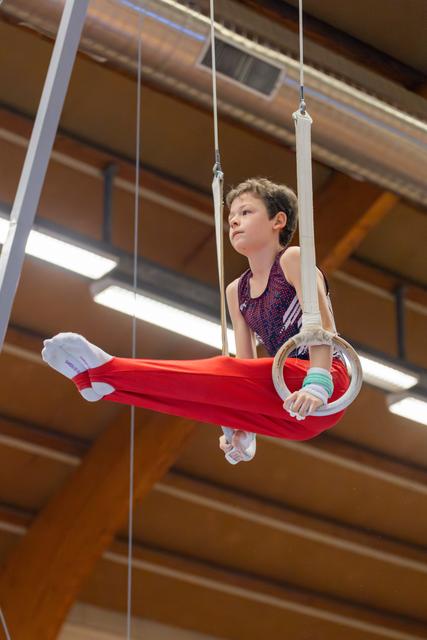 Young boy performs on gymnastics rings with focused expression, legs extended, wearing red pants in a wood-beamed gym.