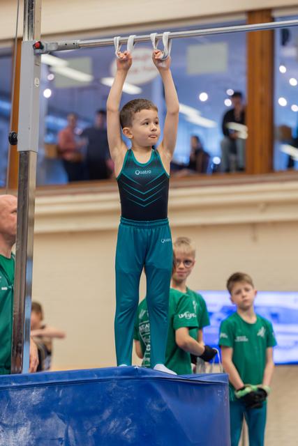 Young boy stands tall on a mat, gripping gymnastics rings overhead with calm focus, teammates watching in the background.