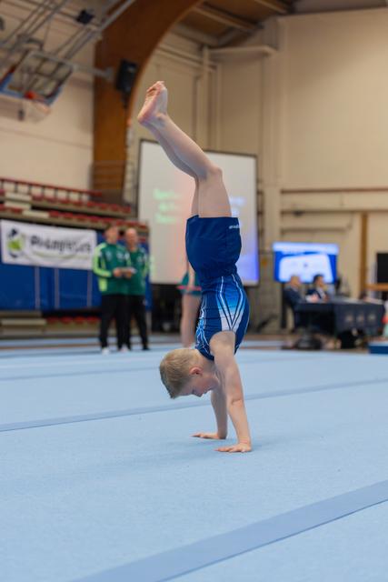 Young male gymnast performing a precise handstand on a blue mat during an indoor gymnastics competition.