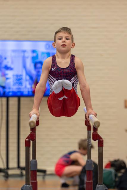 Young male gymnast holds a straddle position on parallel bars, focused expression, wearing red shorts and GK leotard.