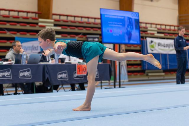 Young boy balances on one leg in an arabesque pose on the floor exercise mat, focused and controlled at a gymnastics meet.