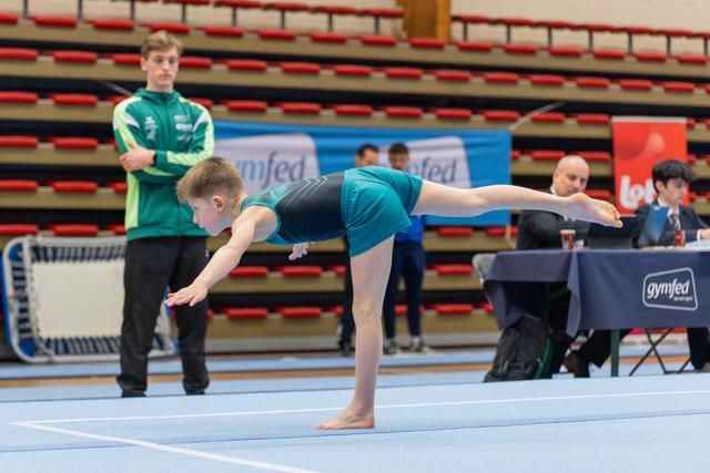 Young boy in teal leotard performs a precise arabesque on the floor mat while a coach in green watches intently.
