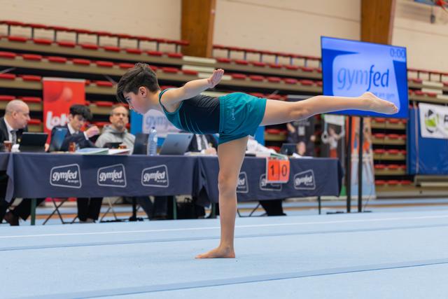 Young gymnast in teal leotard performs a focused arabesque on floor, judges' table visible behind her.