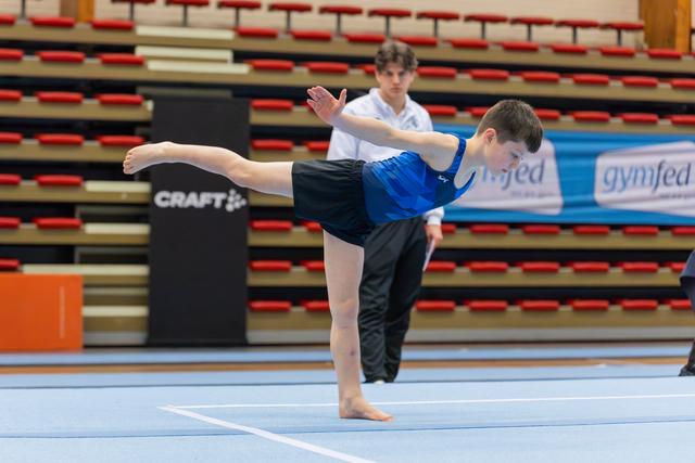 Young male gymnast holds a precise arabesque on the floor, arms extended, coach watching attentively in background.
