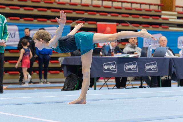 Young gymnast holds a precise arabesque balance on the floor, arms raised, leg extended, focused during a Gymfed event.