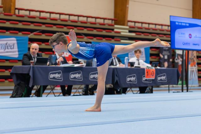 Young boy with glasses performs a focused arabesque on the floor, judges seated at gymfed table behind him.