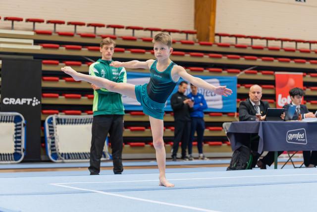 Young gymnast performs a precise arabesque on floor, arms spread wide, focused expression, judges table visible behind.