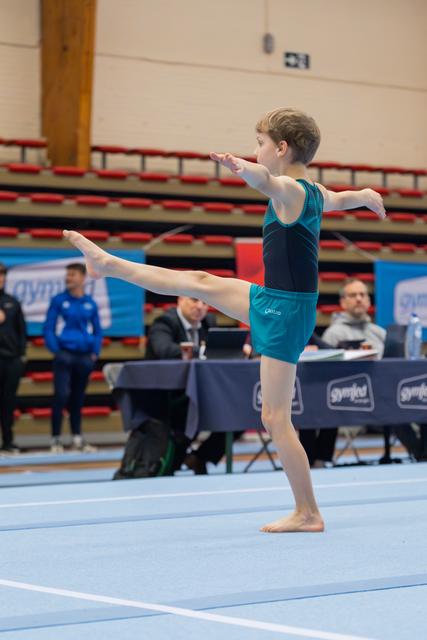 Young male gymnast performs a precise arabesque on the floor, arms outstretched, judges watching at table behind.