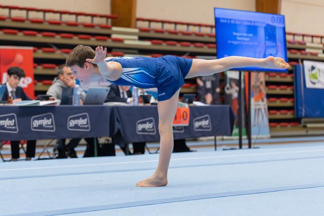 Young gymnast in blue leotard holds a perfect arabesque balance on the floor, judges seated at table behind.