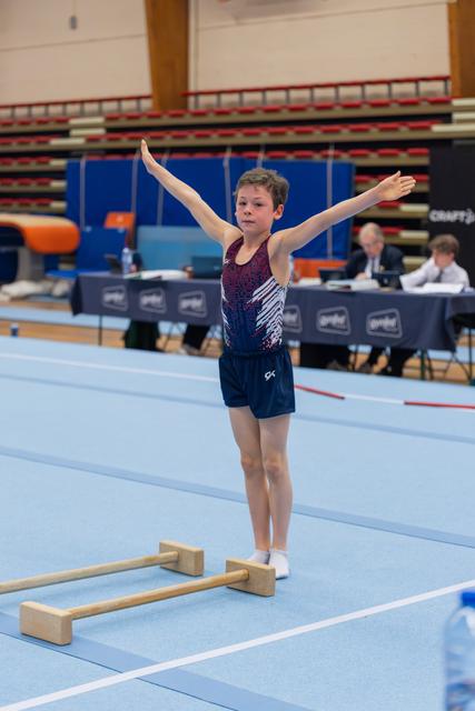 Young boy gymnast stands proudly with arms outstretched on blue floor mat, judges panel visible behind him.