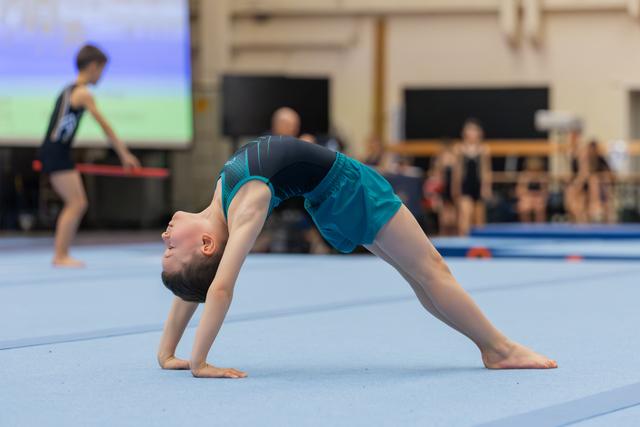 Young gymnast performs a bridge pose on the blue floor mat, head dropped back, focused expression during a gymnastics event.