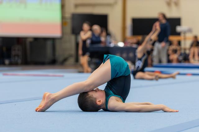 Young gymnast performs a deep backbend on the blue floor mat, feet touching head, showing impressive flexibility.