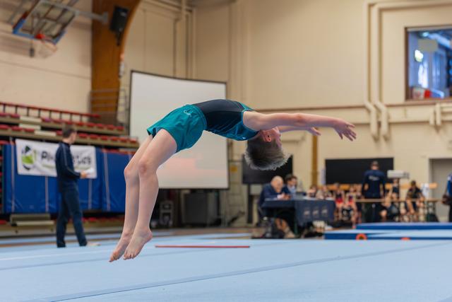 Young gymnast mid-backflip in teal leotard, arched elegantly over the blue floor mat during an indoor gymnastics meet.